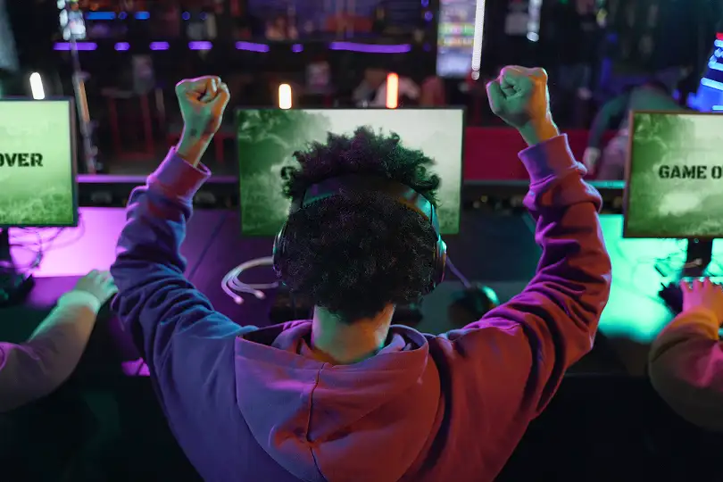Rear view of a gamer wearing headphones raising both fists in celebration in a purple-lit gaming room
